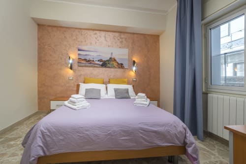 Camera da letto moderna di Chronocasa Messina con biancheria viola, parete in sughero, cuscini gialli e faro costiero