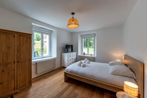 Chambre spacieuse au Refuge des Brumes avec lit en bois, draps blancs, commode TV, penderie et vue montagne