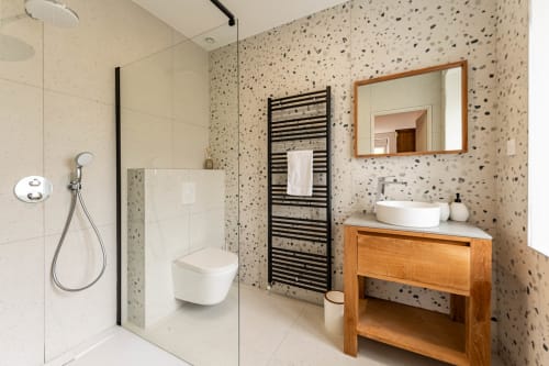 Salle de bains moderne au Refuge des Brumes avec murs en terrazzo, douche à l'italienne, porte-serviettes noir, vasque en bois et miroir