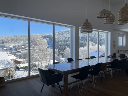 Salle à manger contemporaine au Refuge des Brumes avec vue sur la forêt enneigée, chaises noires et suspensions en fibres tressées