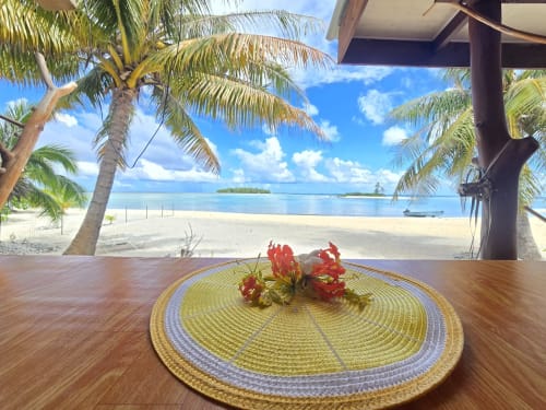 Table en bord de plage au Tikehau Herearii avec fleurs rouges, palmiers et vue sur le lagon turquoise à Tuherahera