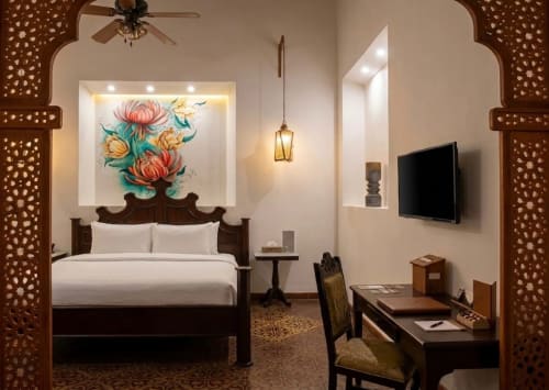 Boutique hotel bedroom in Merida with dark wood bed, white linens, floral artwork, flat-screen TV, desk, and ornate wooden