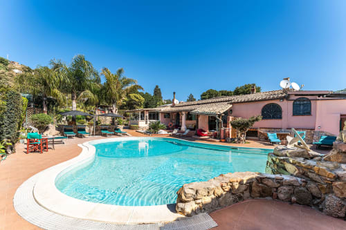 Piscina turchese con bordo bianco a Villa Verde, villa rosa e palme sotto un cielo azzurro limpido