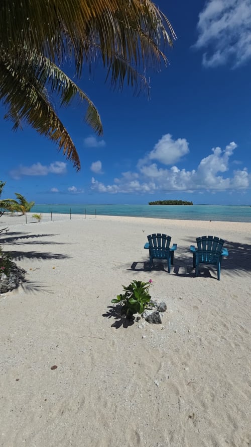 Deux chaises Adirondack bleu ciel sur le sable blanc de la plage de Tikehau Herearii, avec lagon turquoise et palmes au-dessus