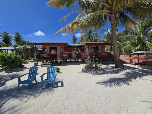 Tikehau Herearii beachfront cottage with red bungalow, blue Adirondack chairs on sand, palm trees, and clear sky in