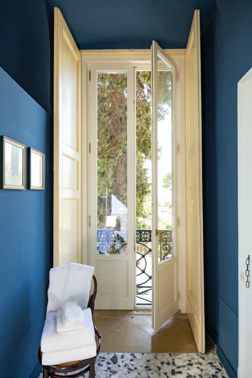 Camera da letto luminosa a Villa Ceci con pareti blu, porte color crema che si affacciano sulla terrazza con vista sul giardino e alberi, sedia bianca, marmo