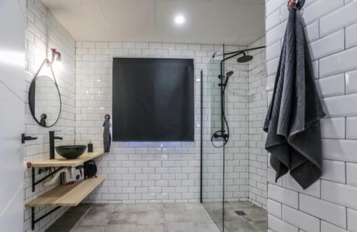 Baño moderno con azulejos subway blancos, accesorios negros, estantes de madera, espejo redondo y ducha abierta en Gestyp-AT