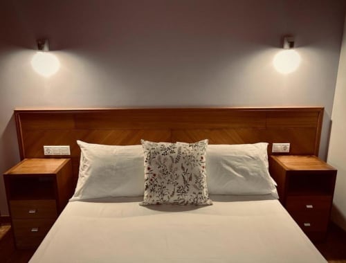 Dormitorio moderno con cabecera de madera, ropa de cama blanca, almohada decorativa y mesitas en Casa Rural A Bouciña