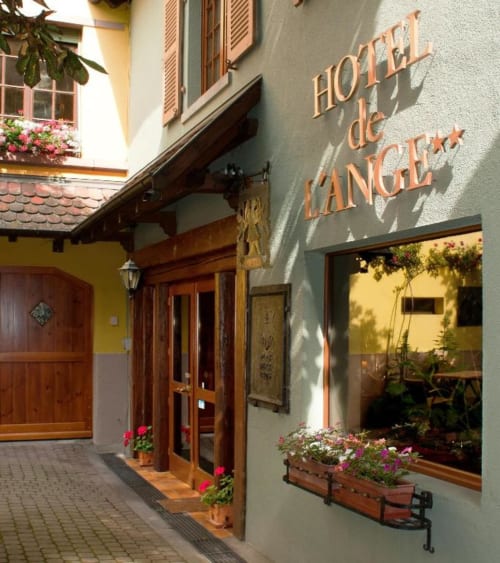 Entrée de cour de l'Hôtel de l'Ange à Niedermorschwihr avec façade en pierre, portes en bois et fleurs roses