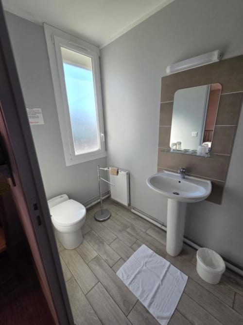 Salle de bains moderne de l'hôtel Le Bel Air avec lavabo blanc sur piédestal, toilettes, miroir, fenêtre vue ville et carrelage