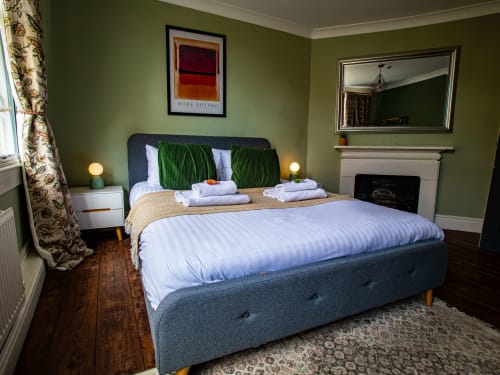 Spacious bedroom at Stonegate Townhouse York with gray upholstered bed, green walls, fireplace, and hardwood flooring.