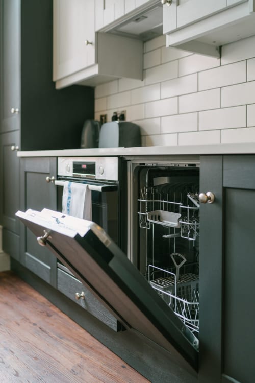 Modern kitchen with dark green cabinetry, white subway tile backsplash, stainless steel appliances and open dishwasher at