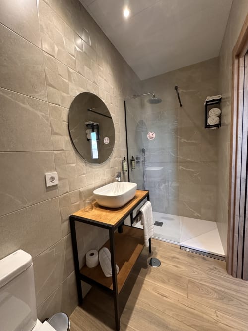 Baño moderno del Hotel Síbel Baza con espejo redondo, lavamanos tipo vasija, ducha abierta y pisos de madera
