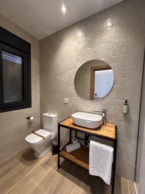 Baño moderno del Hotel Síbel Baza con lavabo blanco, espejo redondo, inodoro y tocador de madera con marco negro