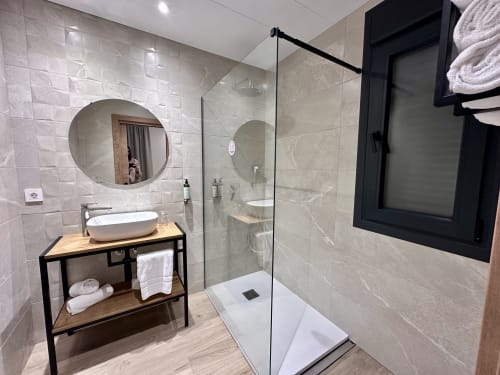 Baño moderno del Hotel Síbel Baza con ducha abierta, espejo redondo, lavamanos blanco y tocador de madera