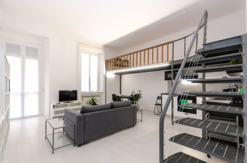 Moderno loft apartment a Milano con divano grigio, letto in mezzanino, scala in metallo, TV e luce naturale dalle finestre