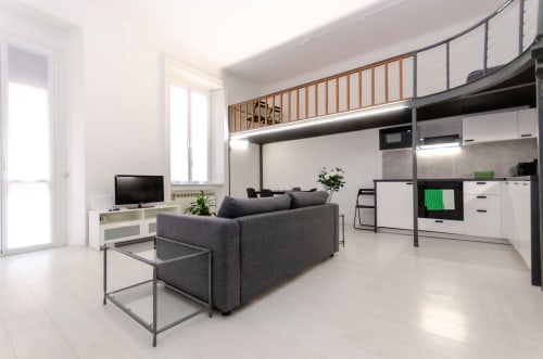 Monolocale moderno a Milano con letto rialzato, divano grigio, TV, angolo cottura e ringhiere in metallo presso Bronzetti Home