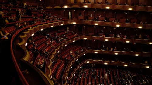 Grand teatro dell'opera con più livelli di balconate rivestite in velluto rosso e illuminazione calda, con capienza per centinaia di ospiti