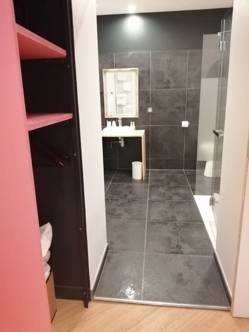Salle de bains accessible à l'Hôtel Du Cygne Tours : douche accessible, lavabo blanc, miroir, carrelage foncé, étagères de rangement rose.