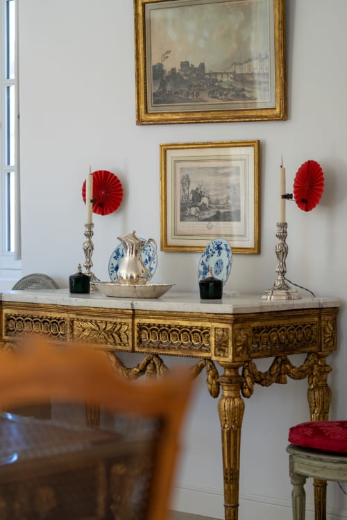 Consola dorada ornamental con tablero de mármol, platos azules y blancos, velas rojas y grabados enmarcados en Le Domaine du Fortin