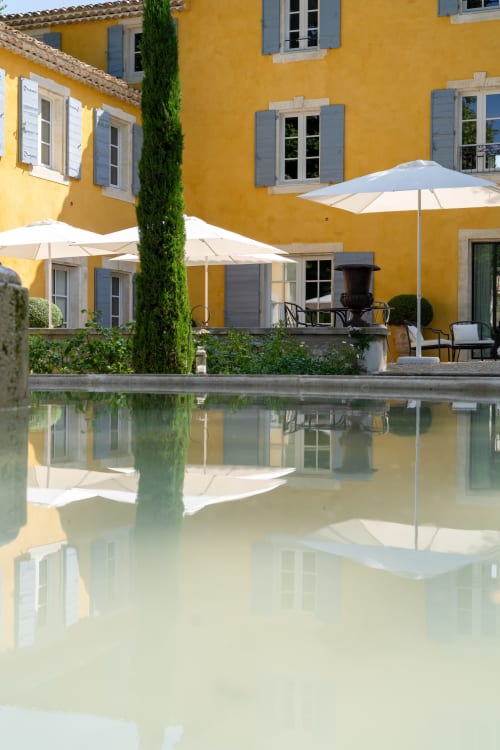 Patio del Domaine du Fortin en Ménerbes con edificio amarillo, espejo de agua, ciprés y sombrillas blancas