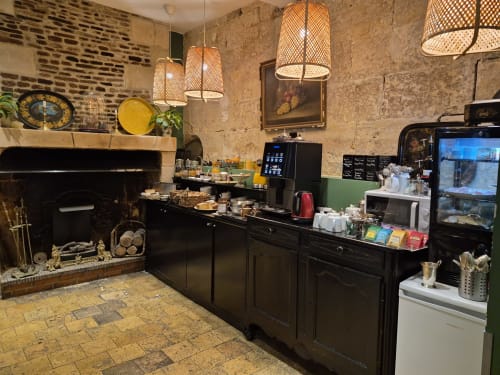 Intérieur rustique du café de l'Hôtel Du Cygne à Tours avec murs en pierre apparente, suspensions tressées, comptoir noir et cheminée.