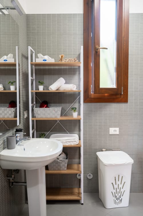 Modern villa bathroom in Ferrara with white pedestal sink, toilet, tiled walls, wooden shelving with towels and toiletries,