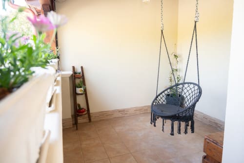 Bright coastal villa room with hanging macramé chair, potted flowers on windowsill, wooden flooring, and cream walls near