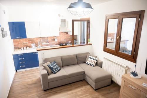 Modern open-plan villa living room with gray sofa, blue kitchen cabinets, brick backsplash, and wooden doors near Ferrara