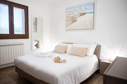 Bright modern bedroom with white linens, beige pillows, wooden furniture, beach scene artwork, and sea view window in Ferrara
