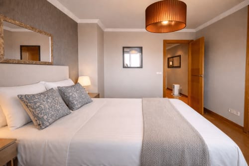 Luminosa habitación con ropa de cama blanca, almohadas grises estampadas, muebles de madera y vista al mar en Holibai de Baiona