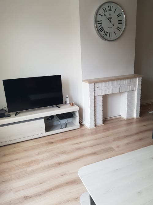 Séjour moderne avec télévision écran plat, cheminée blanche, sol en bois et horloge murale à la Résidence malouine