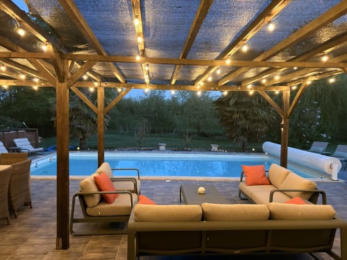Élégant espace de détente au bord de la piscine du Jardin d'Elusa à Eauze, avec pergola illuminée, sièges confortables et eau bleue scintillante