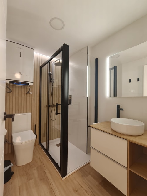 Baño moderno en Bahia Mar Canarias con ducha de marco negro, lavamanos blanco y tocador de madera
