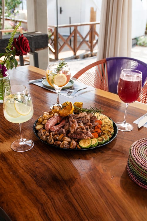 Mesa de comedor al aire libre en Meraki Glampings con carnes a la parrilla, verduras asadas y cócteles coloridos con vistas a Malinalco