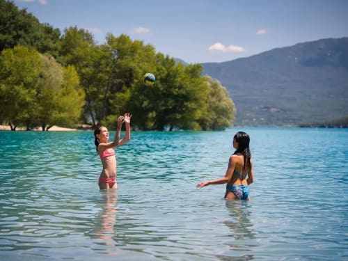 Deux enfants jouent au volley dans les eaux cristallines du lac près des Insolites de la Bastide, entourés de forêts verdoyantes et de montagnes