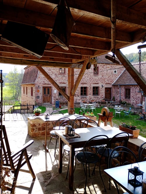 Cour de la Paulusmühle à Soucht : patio couvert avec tables, chaises, bâtisses en pierre, poutres bois et jardins luxuriants
