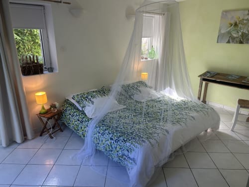 Chambre lumineuse avec lit double, voile de baldaquin, linge de lit motifs verts, carrelage et vue sur jardin