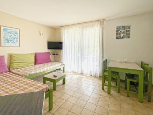 Séjour lumineux au Vallon des Sources avec mobilier vert, coussins roses et jaunes, table à manger, téléviseur