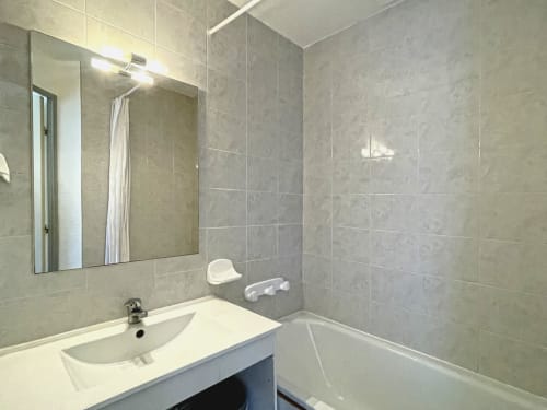 Salle de bain moderne de l'appartement Le Vallon des Sources avec carrelage gris, lavabo, miroir éclairant et baignoire blanche