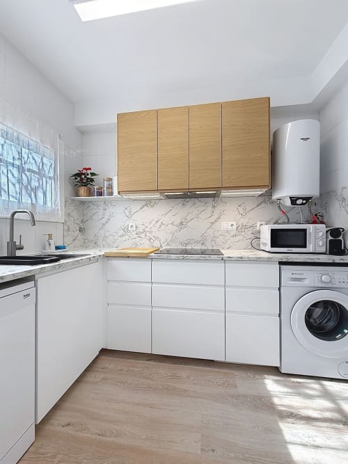 Cocina blanca moderna con armarios superiores de madera, salpicadero de mármol, electrodomésticos de acero inoxidable y lavadora en L'Estartit