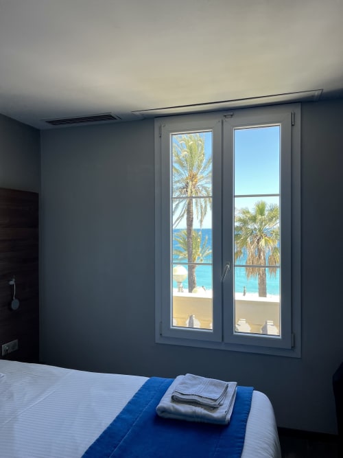 Chambre d'hôtel moderne à l'Hôtel Flots d'Azur Nice avec vue mer, palmiers et plage à travers de grandes fenêtres