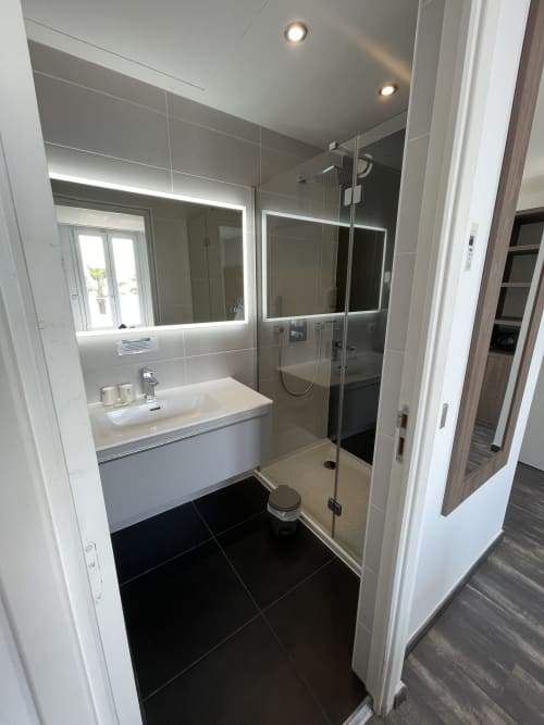 Modern bathroom at Hotel Flots d'Azur Nice with illuminated mirror, white sink, glass shower enclosure, and black tile