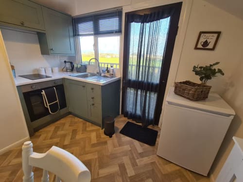 Compact kitchen at 2 Doves Holiday Property in Exeter with sage green cabinets, oven, sink, and white fridge overlooking