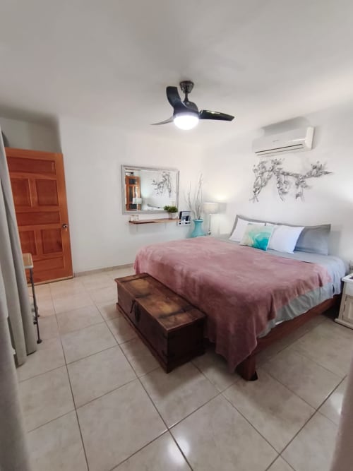 Dormitorio luminoso con cama matrimonial, ropa de cama rosa, ventilador de techo, aire acondicionado, baúl de madera y puerta roja en Casa Punta Perula
