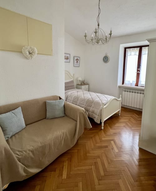 Camera spaziosa con letto singolo, divano beige, pavimento in parquet, lampadario e finestra con persiane rosse a Il Narciso del