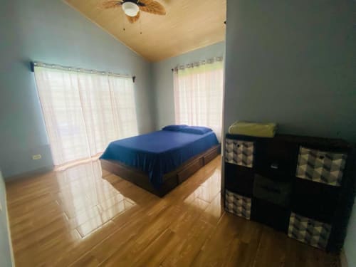 Dormitorio amplio de villa Cabinas Arenal Home con cama azul, pisos de madera, almacenamiento, ventilador de techo y vistas al jardín
