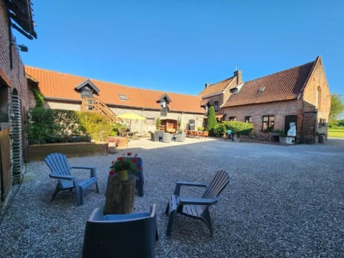 Cour de la Ferme de la Forterie à Verlinghem avec chaises Adirondack bleues, terrasse gravillonnée et bâtiments historiques.