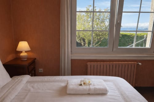 Dormitorio luminoso en la casita MarySol, Baiona con vistas al mar por amplios ventanales, paredes color terracota, ropa de cama blanca y lámpara