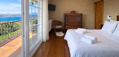 Dormitorio con balcón vista al mar sobre la costa de Baiona, suelo de madera, cama blanca, cómoda de madera y terraza